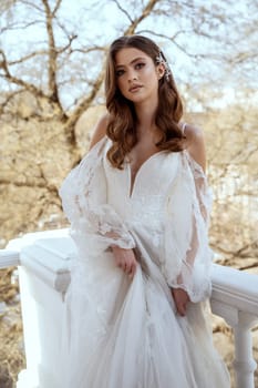 Young brunette bride in traditional long white dress posing on hotel balcony in spring. white dress with long sleeves, corset and open shoulders. Bride portrait, wedding makeup, fashionable bride.