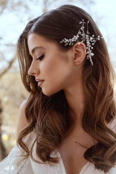 close up of profile of young brunette bride in traditional white dress. Long hair styled in waves, white decoration of small pearls in hair on the side. Portrait of bride, wedding makeup and hairstyle