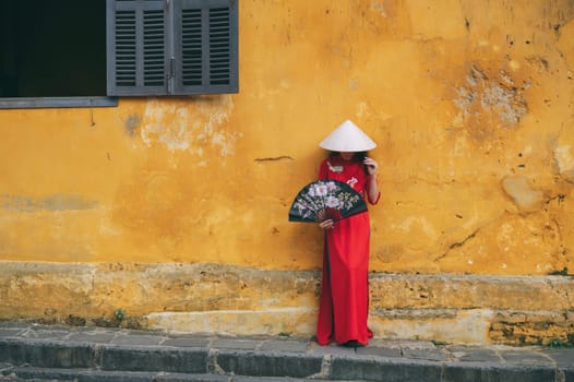 Asian young woman traveler tourist in traditional Vietnamese non la hat and red dress on old town street in Hoi An in Vietnam in Asia