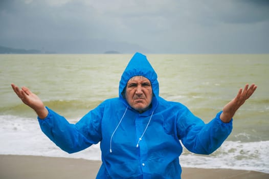 sad elderly man in a raincoat on a beach by the sea with waves in cloudy weather. The concept of the rainy and monsoon season