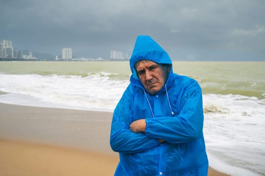 sad elderly man tourist in a raincoat on a beach by the sea with waves in cloudy and rainy weather. Concept of the rainy and monsoon season