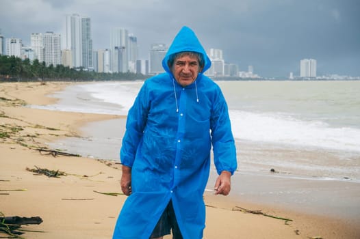 Sad elderly man tourist in a raincoat on a dirty beach by sea after a storm in cloudy weather. The concept of the rainy and monsoon season
