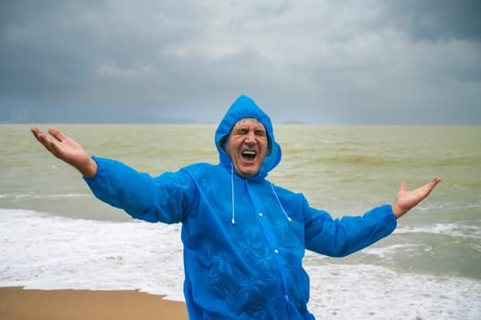 happy emotional elderly man tourist in a raincoat on beach by the sea after a storm in cloudy weather. The concept of rainy and monsoon season