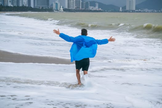 back of a man in a raincoat running along the beach by the sea with waves in cloudy weather with rain. The concept of the rainy and monsoon season