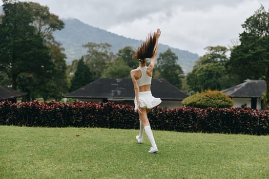 A young woman in a white sports outfit, dancing happily in a vibrant green park with mountains behind, embodying joy and freedom in nature