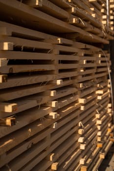 A close-up of neatly stacked wooden boards drying in sunlight at a sawmill. The pattern of the planks creates an industrial texture with rich golden tones.