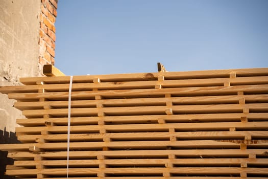 A wide view of fresh timber boards stacked for drying in the sun beside a manufacturing facility. The strong shadows highlight the raw texture of the wood.