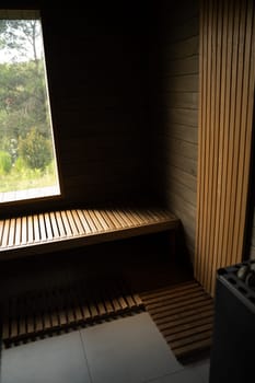 Interior of a wooden sauna with clean slatted bench design and vertical wood paneling. Natural light enters through the window with view of green trees outside.