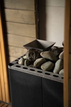 Close view of a metal sauna stove filled with smooth stones, positioned in a wood-paneled relaxation room. Natural light highlights the warm texture and form.
