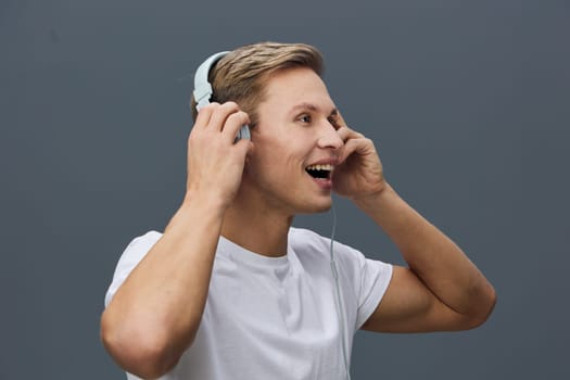 Cheerful young man with light brown hair, headphones on ears, smiling broadly while listening to music, casual white t-shirt, neutral gray background, studio portrait, expressing joy and relaxation, lifestyle and entertainment concept.