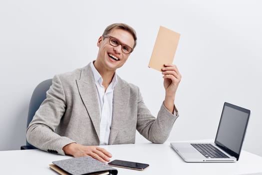 smiling man remote work holding envelope sitting at white desk with laptop and smartphone in casual gray jacket. cheerful young caucasian male professional using digital devices for communication and freelance tasks at home office. Concept: digital nomad and communication