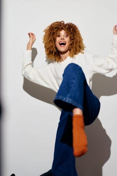 lively young woman expressing joy and energy through dance Dressed casually in a white sweater and jeans, she poses against a bright white background Her curly hair and colorful socks contribute to