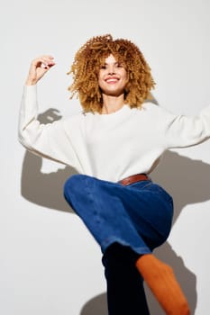 Cheerful woman with voluminous curly hair wearing a cozy white sweater and denim jeans, striking a dynamic pose on a neutral white backdrop, radiating positivity and confidence