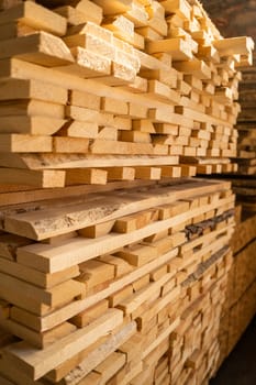 Large pile of pine boards stacked for air drying inside a workshop, lit with strong sunlight. Wood grain, texture, and natural edges clearly visible.