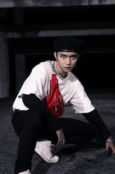 an Asian man sitting around an old building with white clothes and a red bag on his body at night