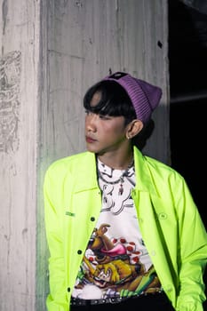 an asian man in a lime colored jacket leaning against a disused building pillar looking very handsome at night