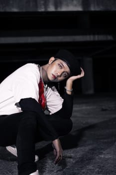 an Asian man sitting around an old building with white clothes and a red bag on his body at night