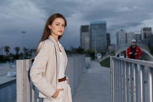 Confident woman in neutral suit poses on urban bridge with city skyline and overcast sky, showcasing fashion style and calm mood in modern outdoor setting