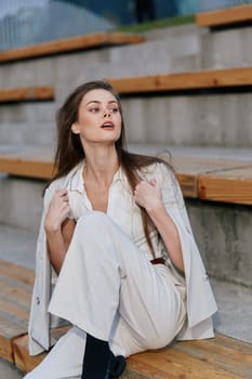 Confident young white woman in elegant white outfit and blazer sits on wooden stairs in neutral gray urban setting, capturing autumn fashion style with calm emotions.