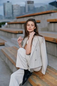 young woman in a light beige suit sits on wooden urban stairs with a thoughtful expression, showcasing fashion style and casual business outfit in a spring city environment