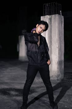 an Asian man dressed all in black and black hair posing as masculine in front of a building pillar