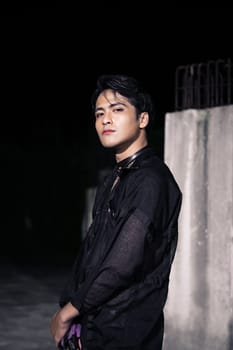 an Asian man dressed all in black and black hair posing as masculine in front of a building pillar