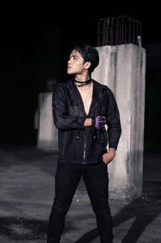 an Asian man dressed all in black and black hair posing as masculine in front of a building pillar