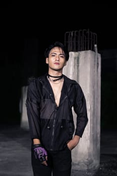 an Asian man dressed all in black and black hair posing as masculine in front of a building pillar