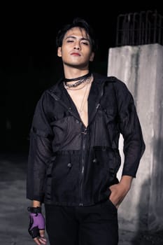 an Asian man dressed all in black and black hair posing as masculine in front of a building pillar