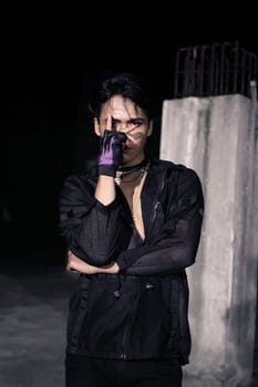 an Asian man dressed all in black and black hair posing as masculine in front of a building pillar