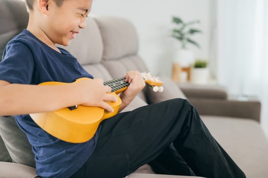 A child's hand plays a yellow ukulele while seated on a sofa in a blue shirt and dark pants. The focus highlights the joy of learning music in a relaxed home setting.