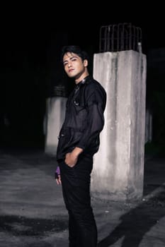 an Asian man dressed all in black and black hair posing as masculine in front of a building pillar