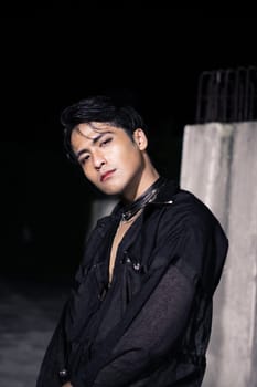 an Asian man dressed all in black and black hair posing as masculine in front of a building pillar