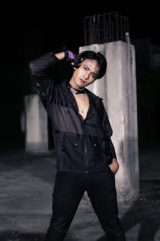 an Asian man dressed all in black and black hair posing as masculine in front of a building pillar