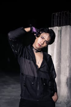 an Asian man dressed all in black and black hair posing as masculine in front of a building pillar
