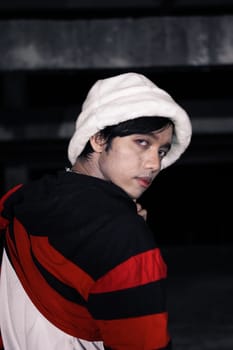 an Asian man in a white velvet hat wears a red sweater and black pants at night