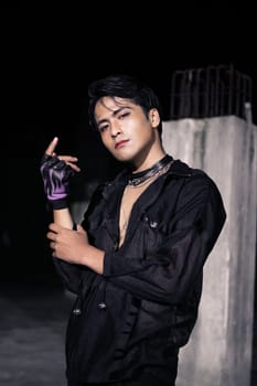 an Asian man dressed all in black and black hair posing as masculine in front of a building pillar