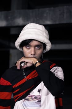 an Asian man in a white velvet hat wears a red sweater and black pants at night
