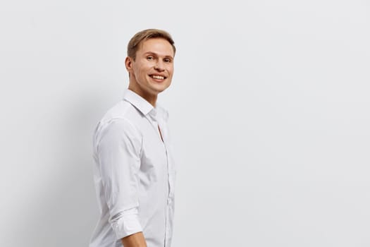 Happy young man with blond hair and blue eyes wearing a white casual shirt, smiling warmly and looking directly at the camera in a bright studio setting with a plain white background. Concept: professional confidence and positivity