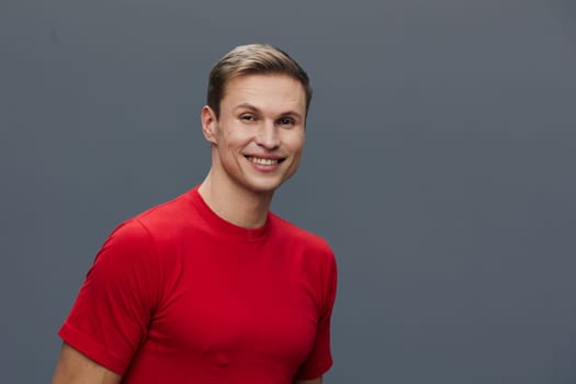 Smiling young caucasian man wear red t-shirt looking directly at camera with friendly expression against gray background in studio portrait style. Concept of virtual communication with digital devices and mental health.