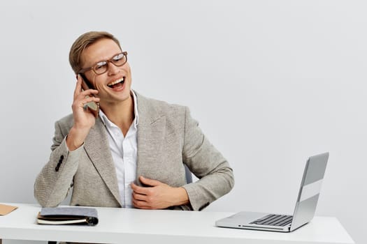 cheerful young man wearing glasses and light gray blazer talking on smartphone and smiling while sitting at white desk with laptop in bright modern space representing digital nomad lifestyle and remote work concept