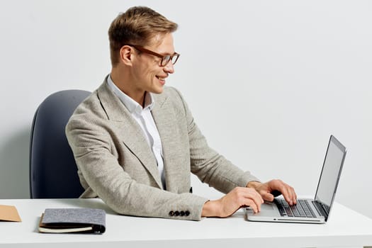Cheerful man with glasses typing on a silver laptop at a clean white desk in a contemporary office, wearing a light gray blazer and white shirt, enjoying work, professional atmosphere, technology, productivity, modern workspace, success, remote working environment.