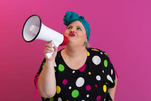 Confident woman in a colorful polka dot outfit and retro headwrap yelling into a megaphone, expressing strong emotion and attitude against a bold pink background.