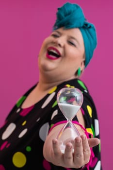 Portrait of a confident woman with a retro headwrap and colorful outfit, holding an hourglass in front of her face as a symbol of time and focus, with a vibrant pink background.