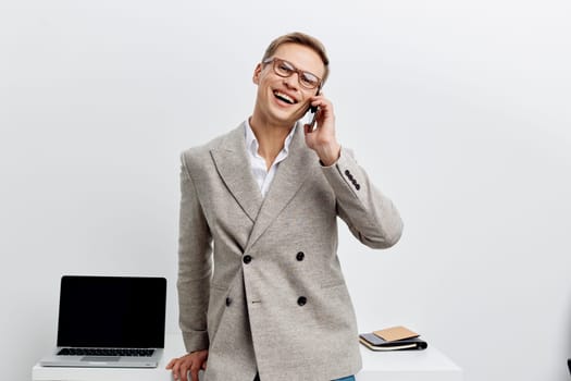 Smiling man in professional attire with glasses making a phone call at modern office workplace, standing in front of desk with laptop and notebook, expressing positivity and communication. Concept: business success.