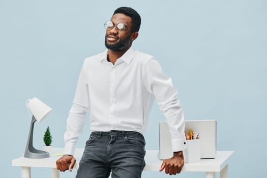 Stylish young Black man dressed in crisp white shirt and dark jeans, posing confidently in a contemporary office setting with a cheerful expression, glasses, and casual professional attire. Bright minimalistic background emphasizes a work and success concept.