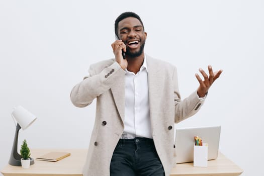 Happy smiling man using mobile phone in office environment, casual business attire, light background with desk, laptop, and stationery, cheerful mood, professional lifestyle scene, concept: communication and success