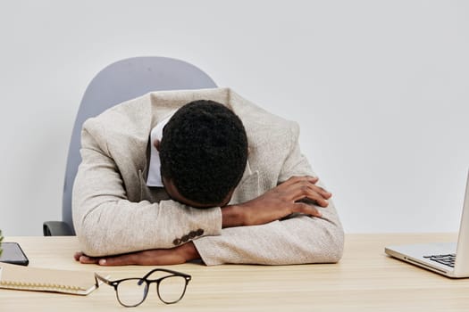 Young adult man in casual office environment with gray background, sitting at a desk with laptop and glasses, resting his head on folded arms, demonstrating fatigue or stress from work, concept of burnout or fatigue, indoor workplace scene