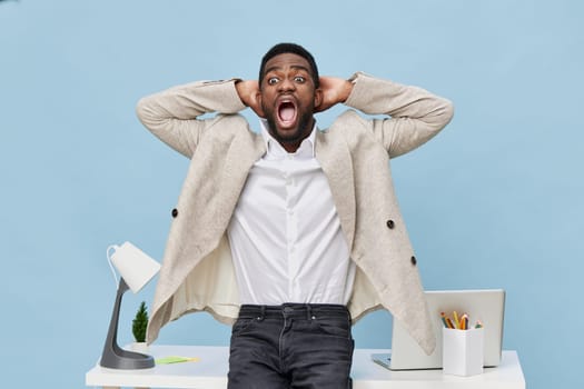 Excited man with youthful appearance, wearing beige blazer and white shirt, expresses shock or frustration at workplace, with hands behind head, in front of cluttered desk, computer, stationery, light blue studio backdrop. Concept: workplace stress and emotions