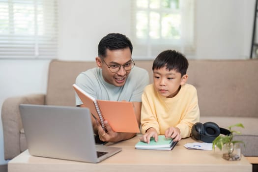 A dedicated father helping his son during homeschool time, reading together from notebooks alongside a laptop.